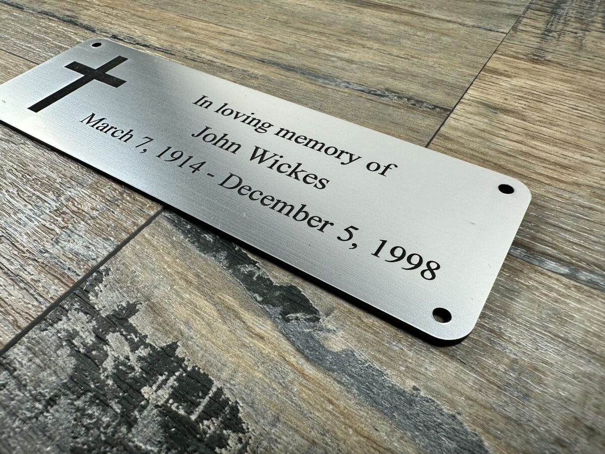 Acrylic Bench Plaque (Cross) Custom 6x2 inch brushed silver acrylic memorial plaque featuring a laser-engraved Christian cross and personalized text for John Wickes (1914-1998). The plaque has a metallic finish and pre-drilled holes, shown resting on a rustic wood surface.