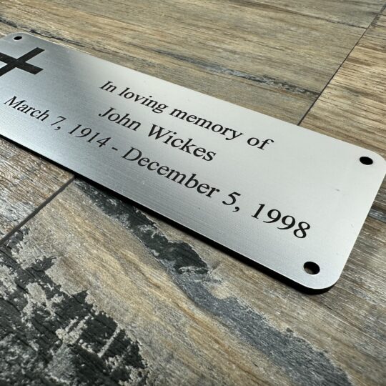 Acrylic Bench Plaque (Cross) Custom 6x2 inch brushed silver acrylic memorial plaque featuring a laser-engraved Christian cross and personalized text for John Wickes (1914-1998). The plaque has a metallic finish and pre-drilled holes, shown resting on a rustic wood surface.