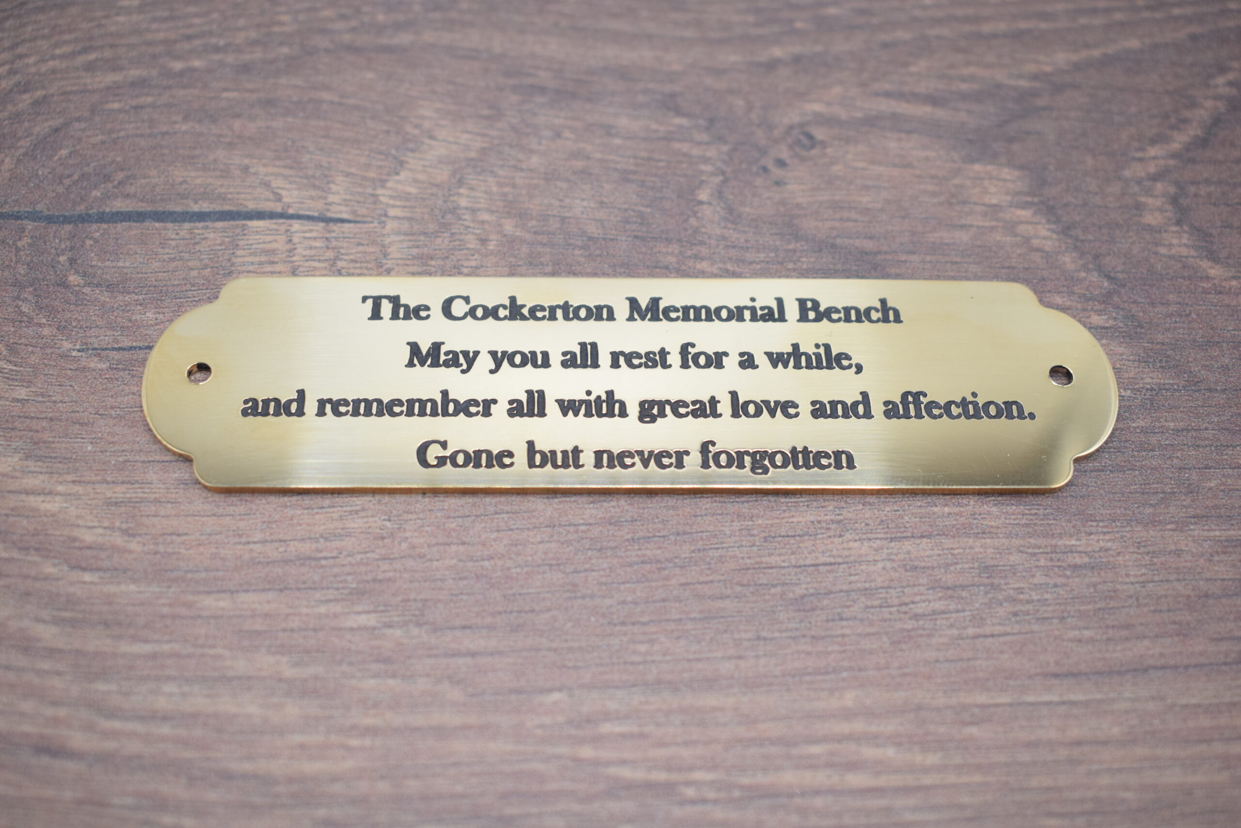Close-up of an engraved brass bench plaque which is 1.5mm thick, featuring dark lettering that reads: "The Cockerton Memorial Bench - May you all rest for a while, and remember all with great love and affection. Gone but never forgotten." It has a decorative scalloped edge and is displayed on a wooden surface