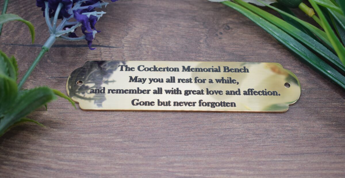 A product photograph of a polished, scalloped brass memorial bench plaque, which is 1.5mm thick. The plaque is engraved with the text, "The Cockerton Memorial Bench - May you all rest for a while, and remember all with great love and affection. Gone but never forgotten." The plaque lies on a dark wood grain surface, flanked by lavender on the left and green foliage on the right.