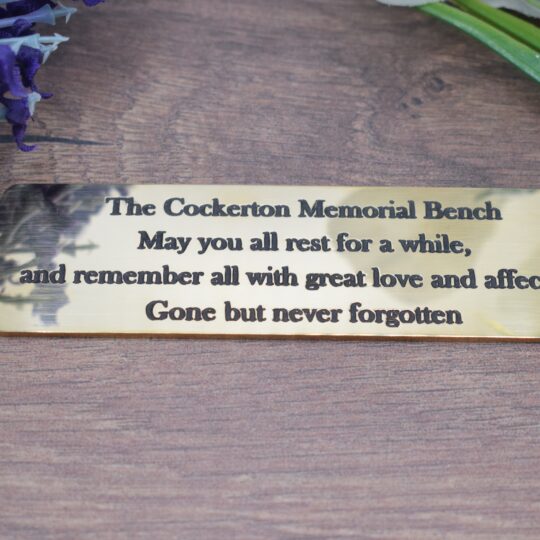 A product photograph of a polished, scalloped brass memorial bench plaque, which is 1.5mm thick. The plaque is engraved with the text, "The Cockerton Memorial Bench - May you all rest for a while, and remember all with great love and affection. Gone but never forgotten." The plaque lies on a dark wood grain surface, flanked by lavender on the left and green foliage on the right.