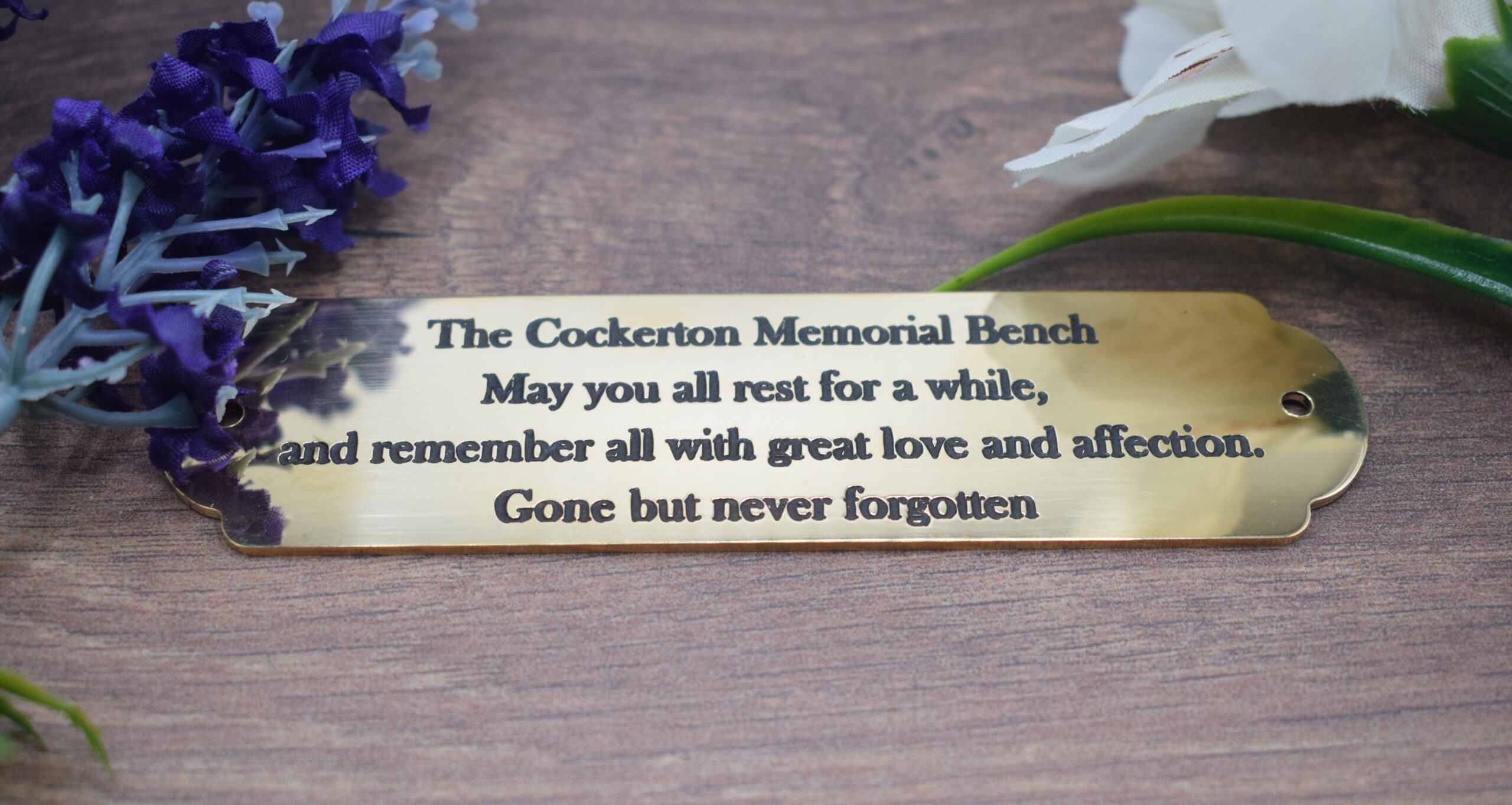 A highly polished 1.5mm thick brass plaque for "The Cockerton Memorial Bench" with dark engraved lettering. The text reads: "The Cockerton Memorial Bench - May you all rest for a while, and remember all with great love and affection. Gone but never forgotten." The plaque features rounded, scalloped ends and pre-drilled holes for mounting.