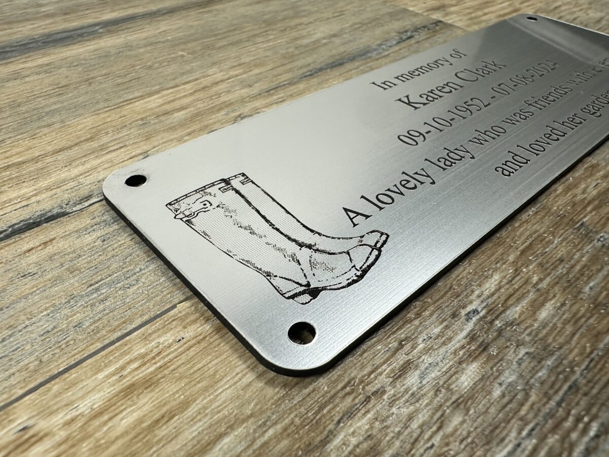 Acrylic Bench Plaque (Wellington Boot) Custom 6x2 inch brushed silver acrylic memorial plaque featuring a laser-engraved wellington boot and text engraved. The plaque has a metallic finish and pre-drilled holes, shown resting on a rustic wood surface.