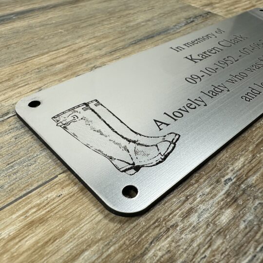 Acrylic Bench Plaque (Wellington Boot) Custom 6x2 inch brushed silver acrylic memorial plaque featuring a laser-engraved wellington boot and text engraved. The plaque has a metallic finish and pre-drilled holes, shown resting on a rustic wood surface.