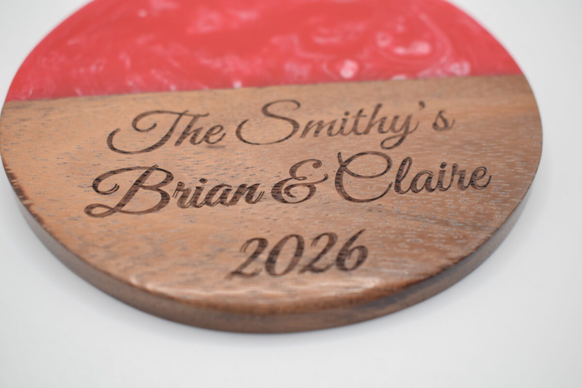 Circular acacia wood and red resin coaster laser-engraved with the cursive text "The Smithy's Brian & Claire 2026".