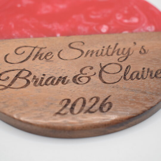 Circular acacia wood and red resin coaster laser-engraved with the cursive text "The Smithy's Brian & Claire 2026".