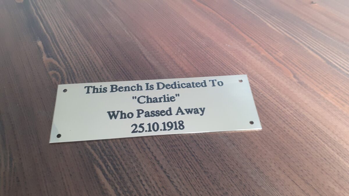 Close-up of an engraved brass bench plaque which is 1.5mm thick, featuring dark lettering that reads: "The Bench is Dedicated - Charlie who passed away 25.10.1918" and is displayed on a wooden surface