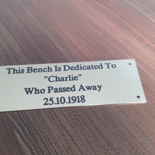 Close-up of an engraved brass bench plaque which is 1.5mm thick, featuring dark lettering that reads: "The Bench is Dedicated - Charlie who passed away 25.10.1918" and is displayed on a wooden surface