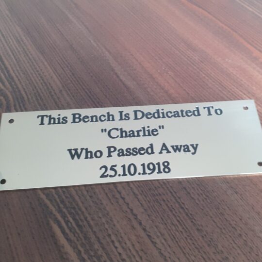 Close-up of an engraved brass bench plaque which is 1.5mm thick, featuring dark lettering that reads: "The Bench is Dedicated - Charlie who passed away 25.10.1918" and is displayed on a wooden surface